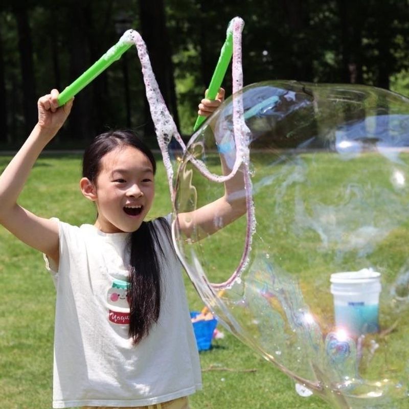 a girl with two wands raised making a giant bubble in a backyard