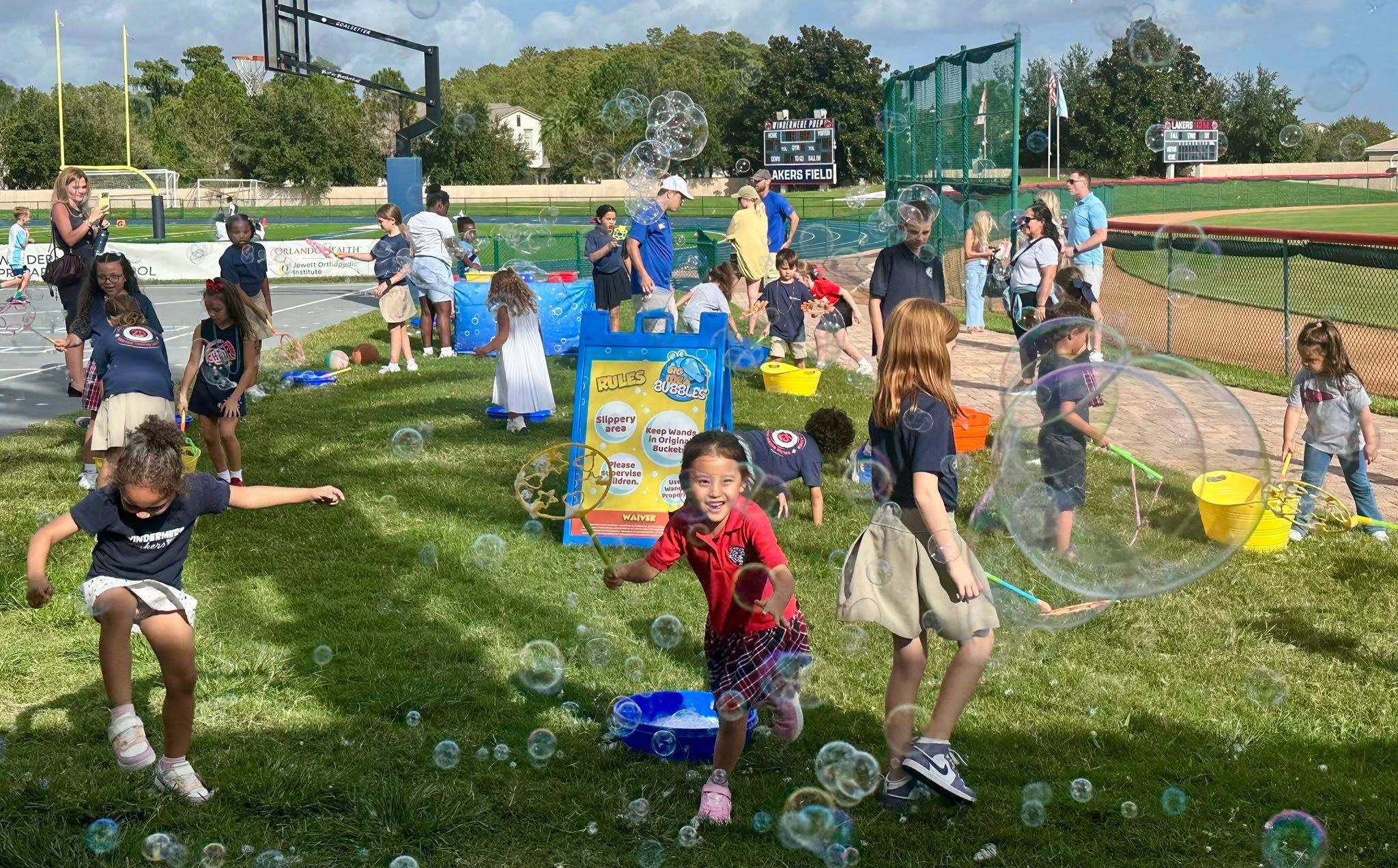 a crowd of kids making giant bubbles beside a Big Idea Bubbles sign at a school field in Orlando