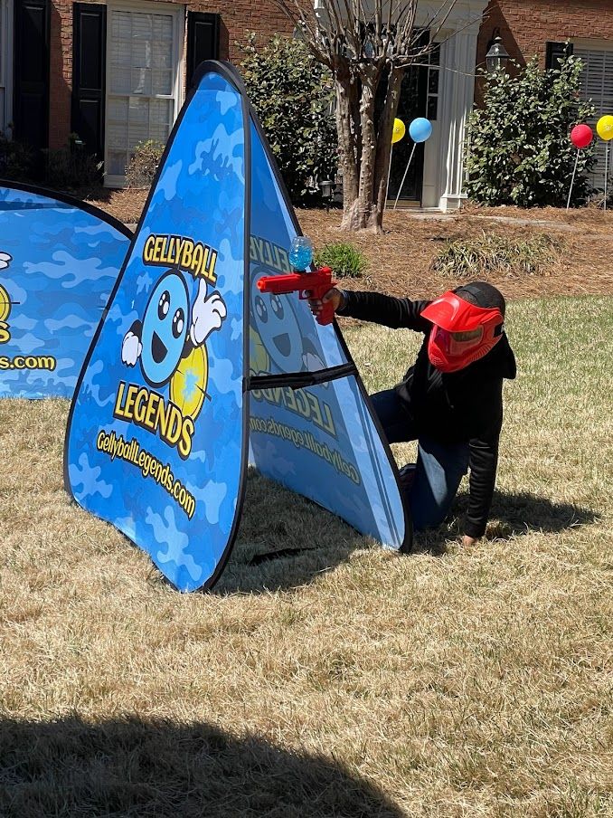 A player in a red mask kneels behind a pop-up bunker on a front lawn with balloons