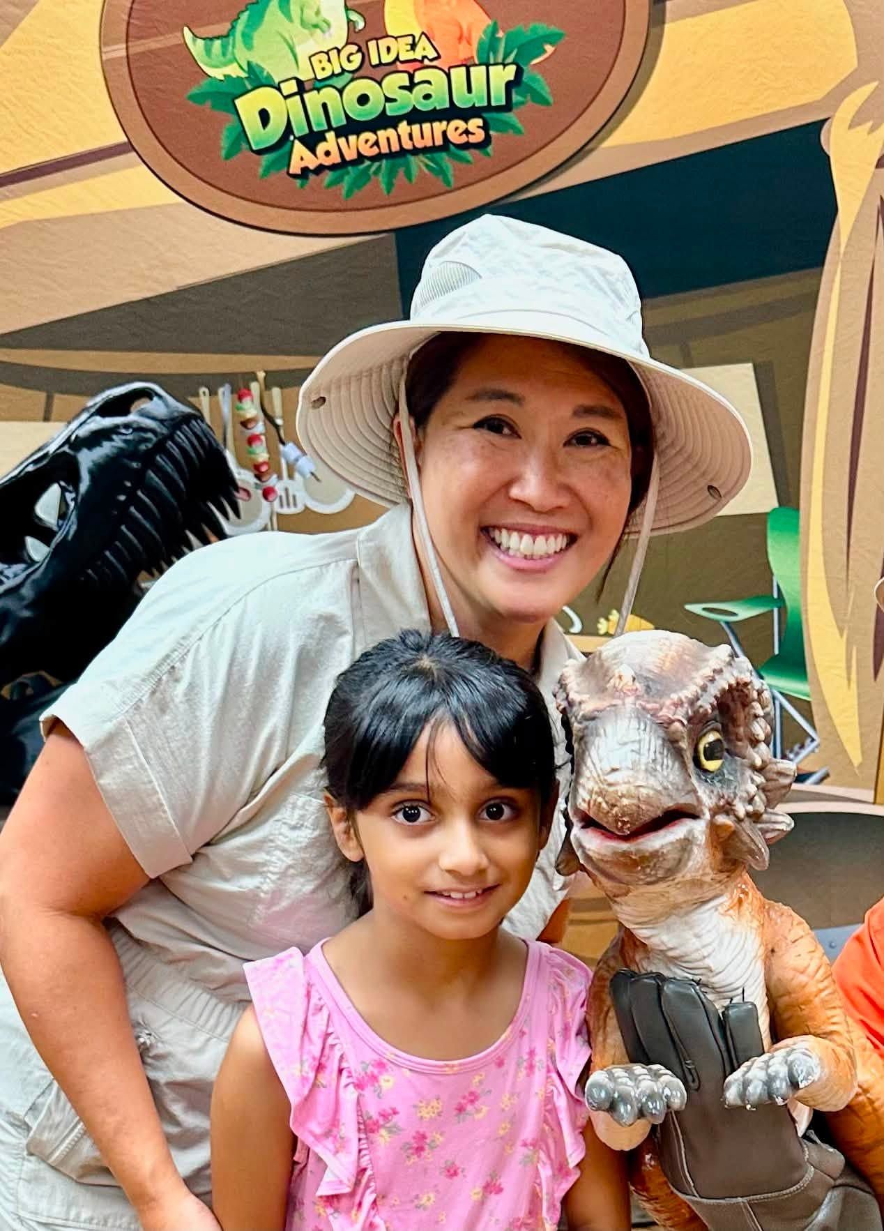 A mom and daughter smile beside a lifelike baby dinosaur with a spiky crest