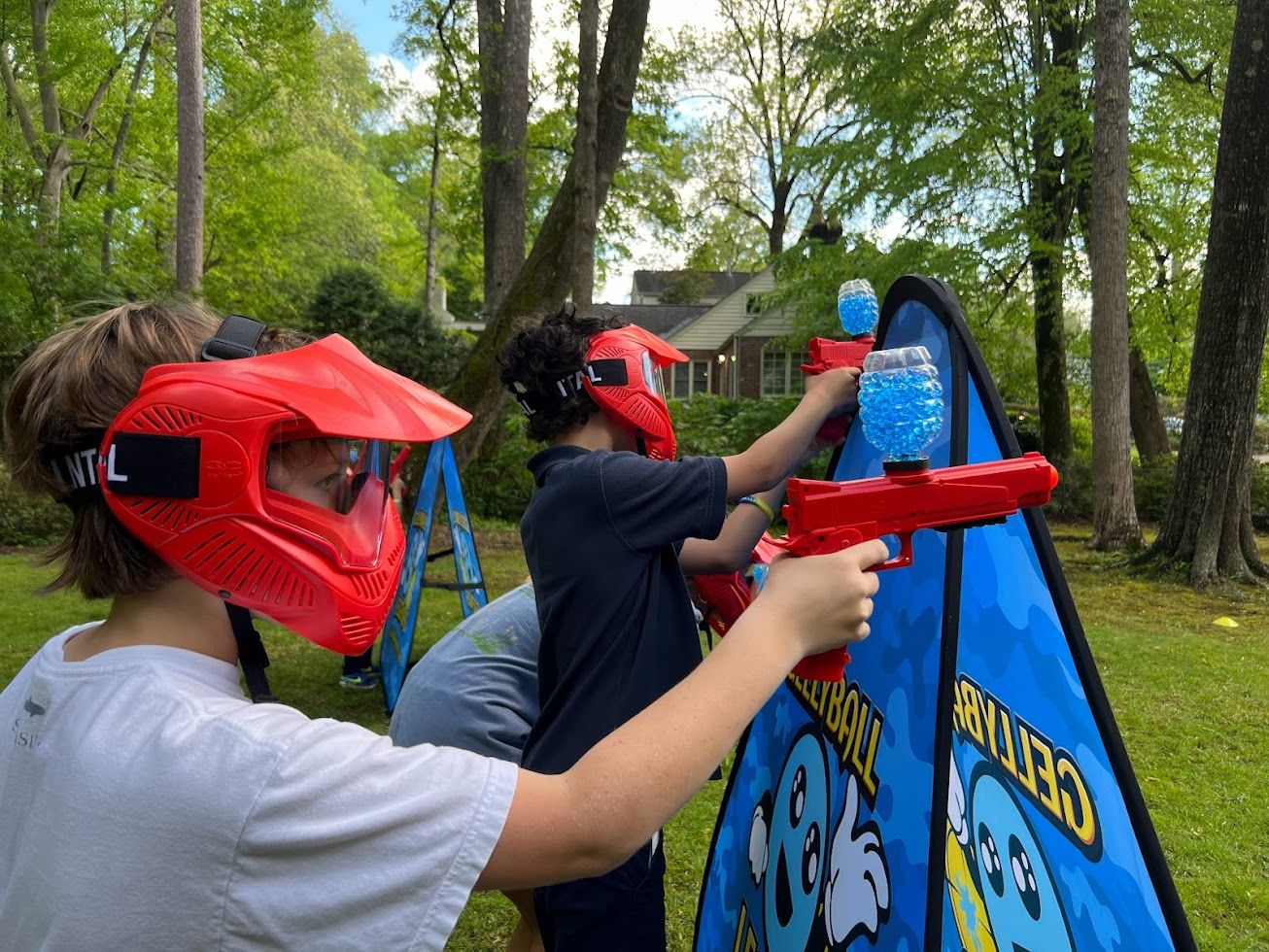 Two players in red masks take aim over a pop-up bunker in a wooded backyard