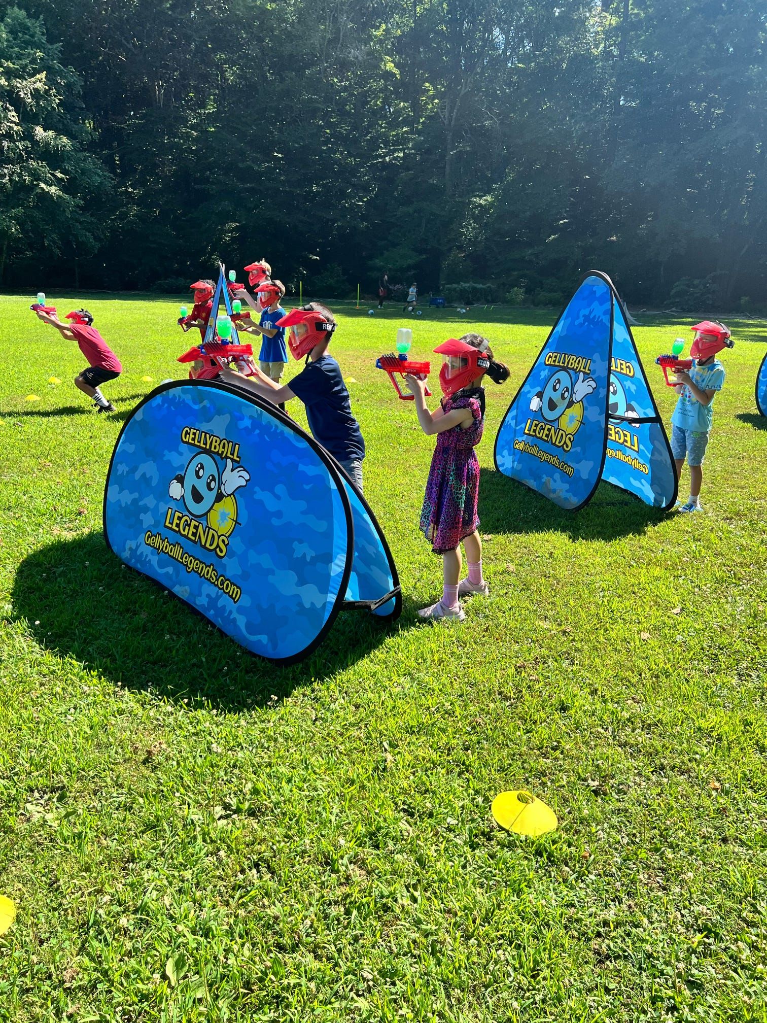 Kids in red masks take aim from behind pop-up bunkers on a sunny park field