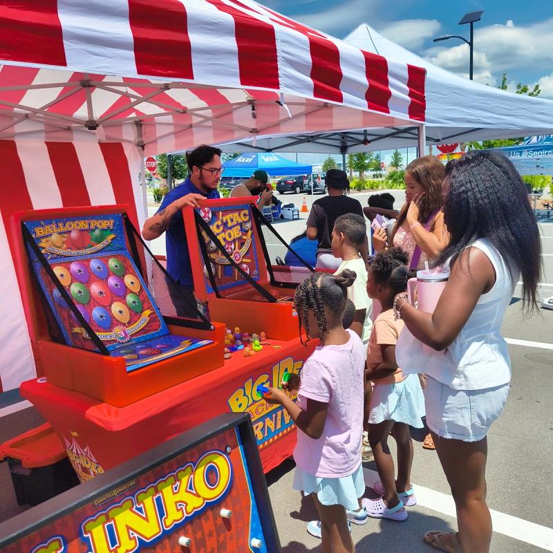kids playing balloon pop and tic-tac-toe while a host runs the booth at an outdoor community event.