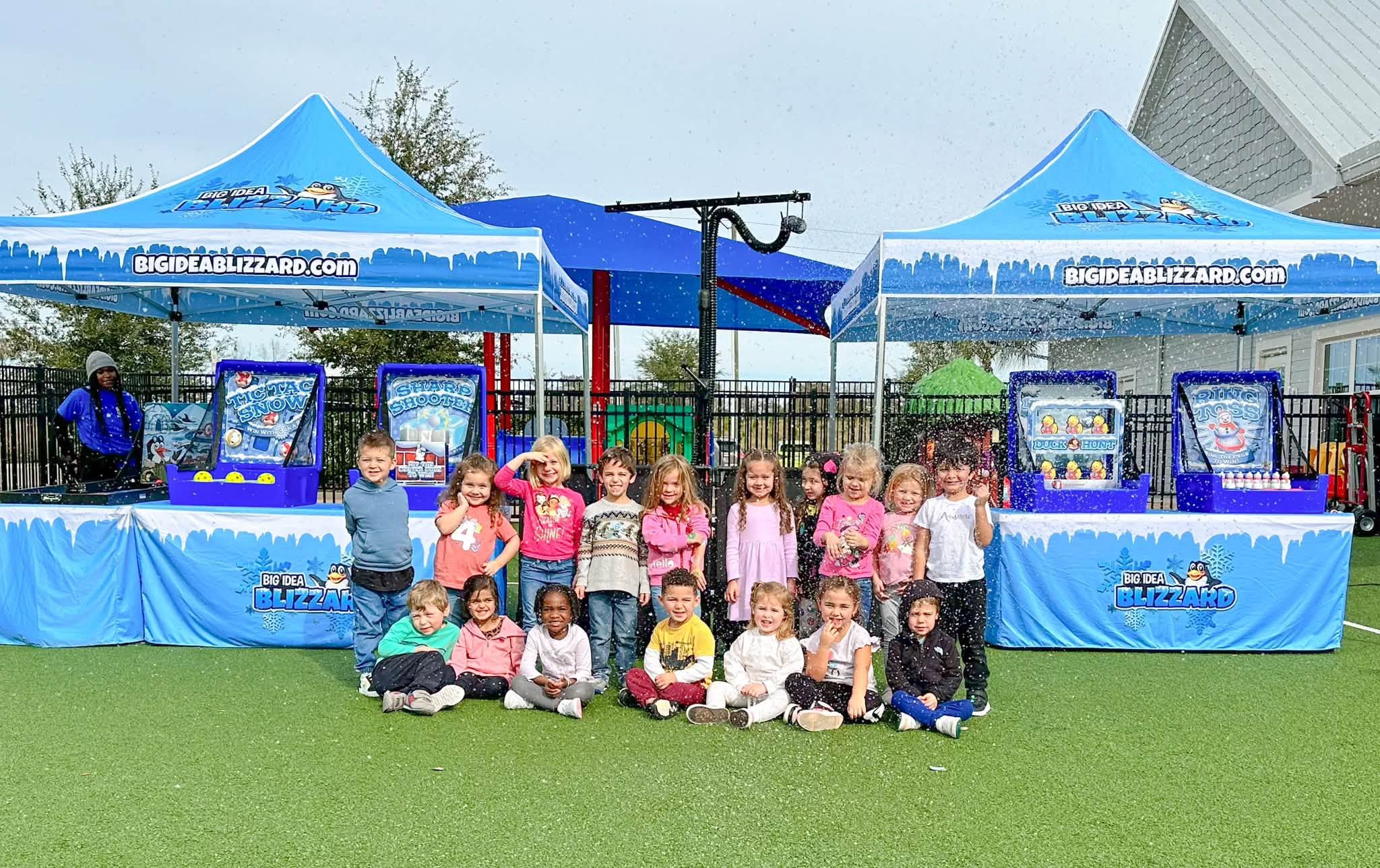 A class of young kids poses on turf in falling snow, with two Blizzard tents and the snow machine behind