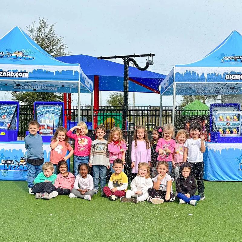 A class of young kids poses on turf in falling snow, with two Blizzard tents and the snow machine behind