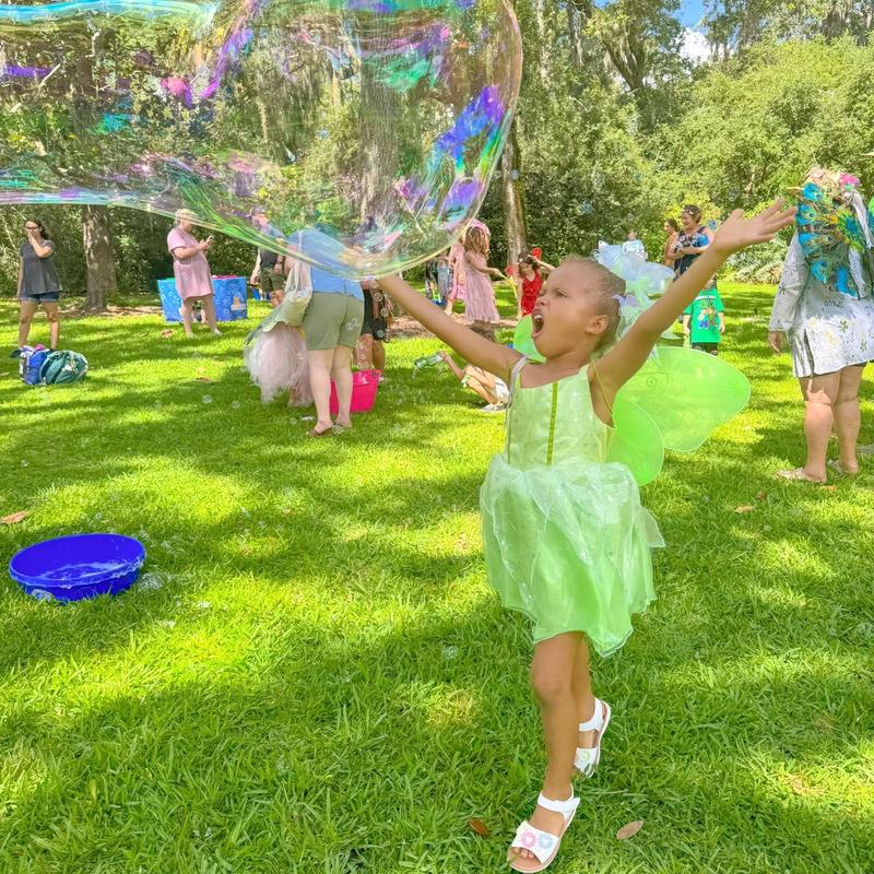 a girl in a green dress reaching up for a giant bubble on a lawn