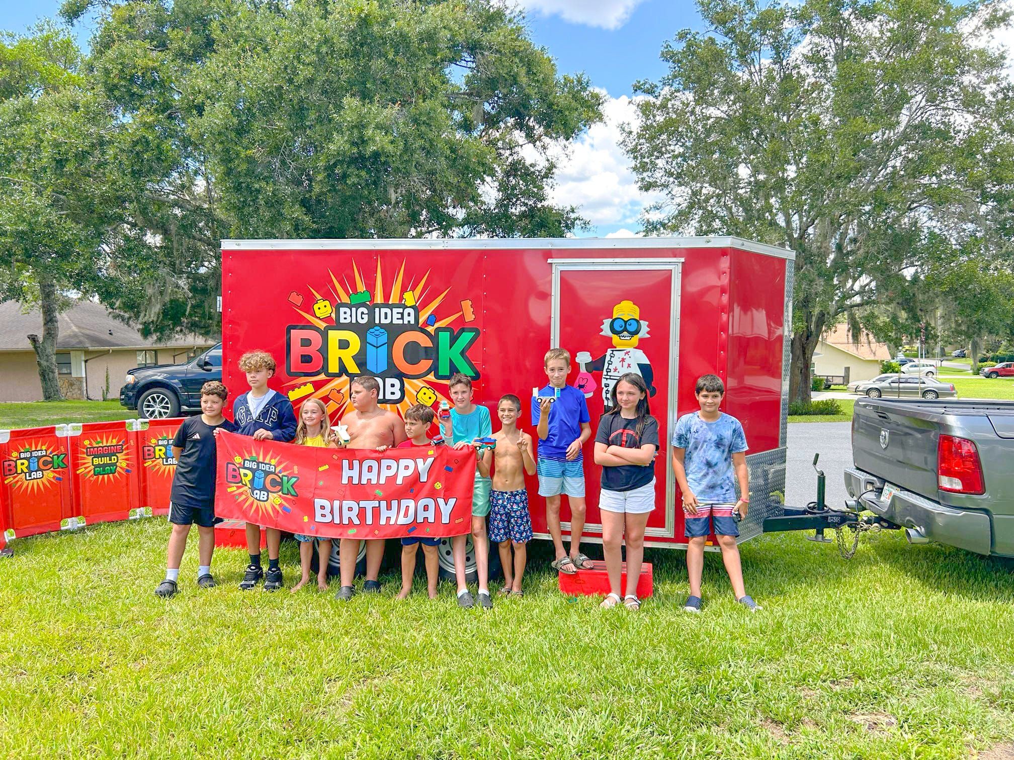 A group of kids holds a Happy Birthday banner in front of the Brick Lab trailer on a shady lawn