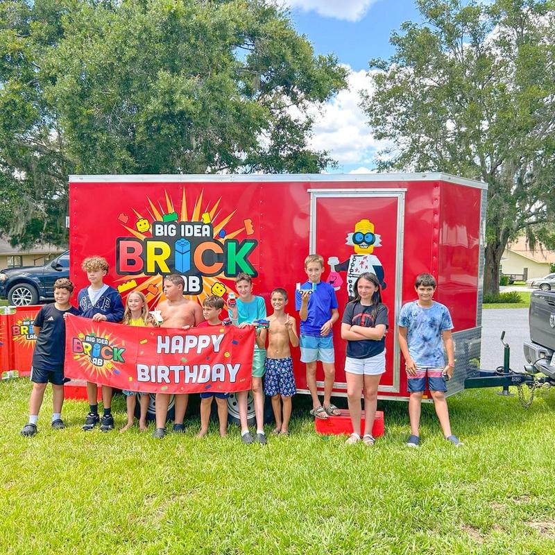 A group of kids holds a Happy Birthday banner in front of the Brick Lab trailer on a shady lawn