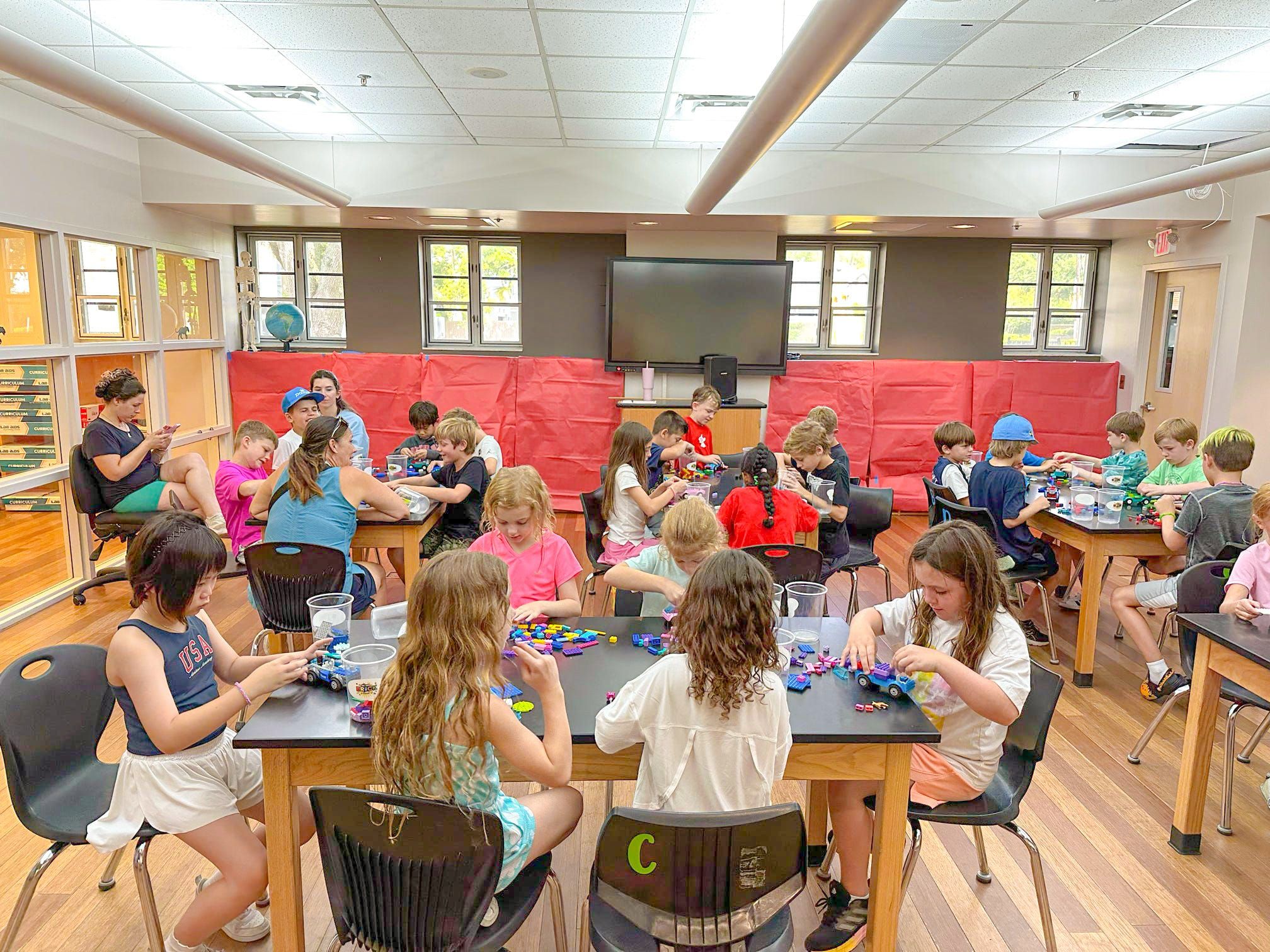 A room full of kids builds with bricks at classroom tables during a Brick Lab session