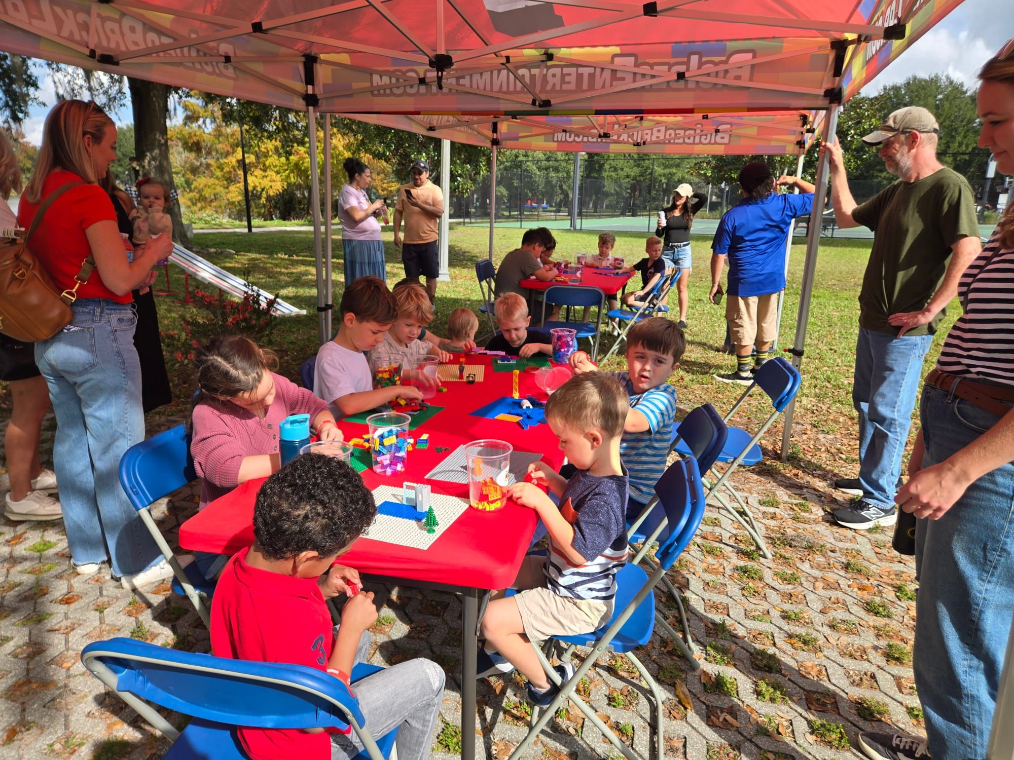 Kids build on baseplates at a red table under a Brick Lab canopy in a park while adults watch