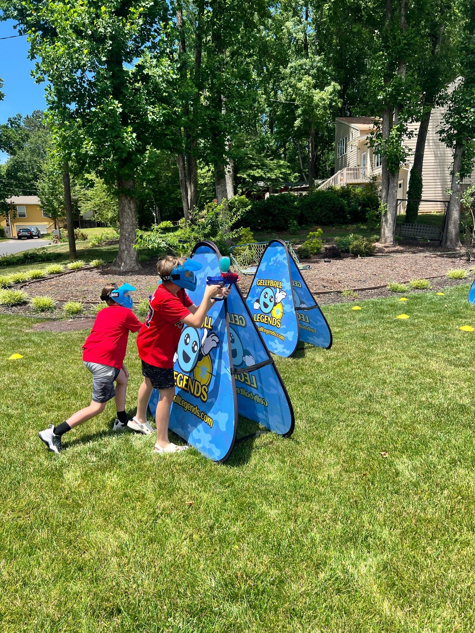 Two boys in red shirts and blue masks take cover behind a bunker in a backyard