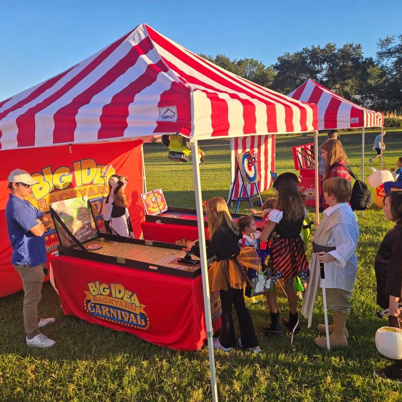 kids lined up to play carnival games under a red and white striped tent at a community event.