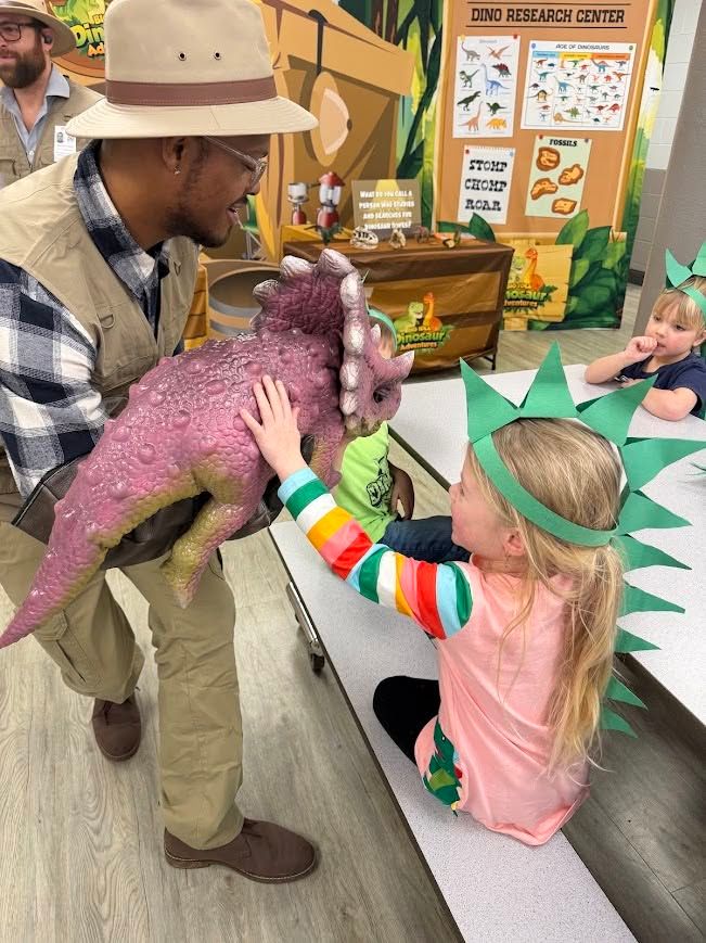 A girl in a paper dinosaur crown pets a pink baby triceratops held by a trainer