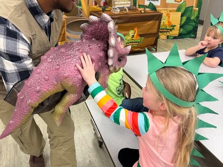 A girl in a paper dinosaur crown pets a pink baby triceratops held by a trainer