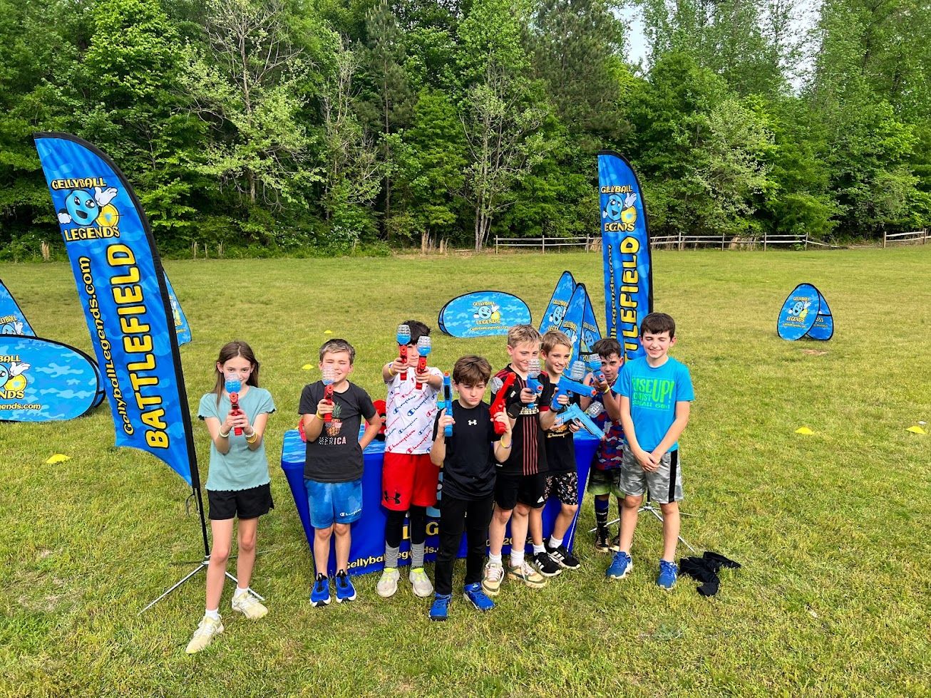 A group of kids hold up their gel blasters in front of feather flags on a big open field