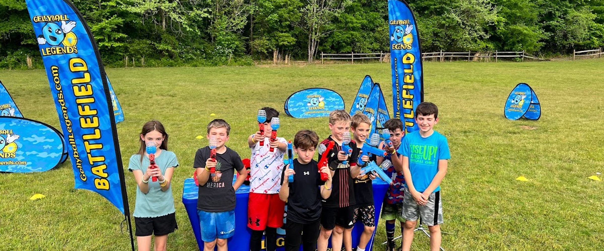 A group of kids hold up their gel blasters in front of feather flags on a big open field