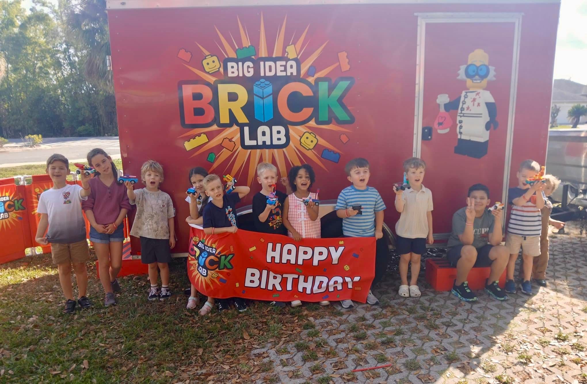 Party guests hold up their brick cars behind a Brick Lab Happy Birthday banner beside the trailer
