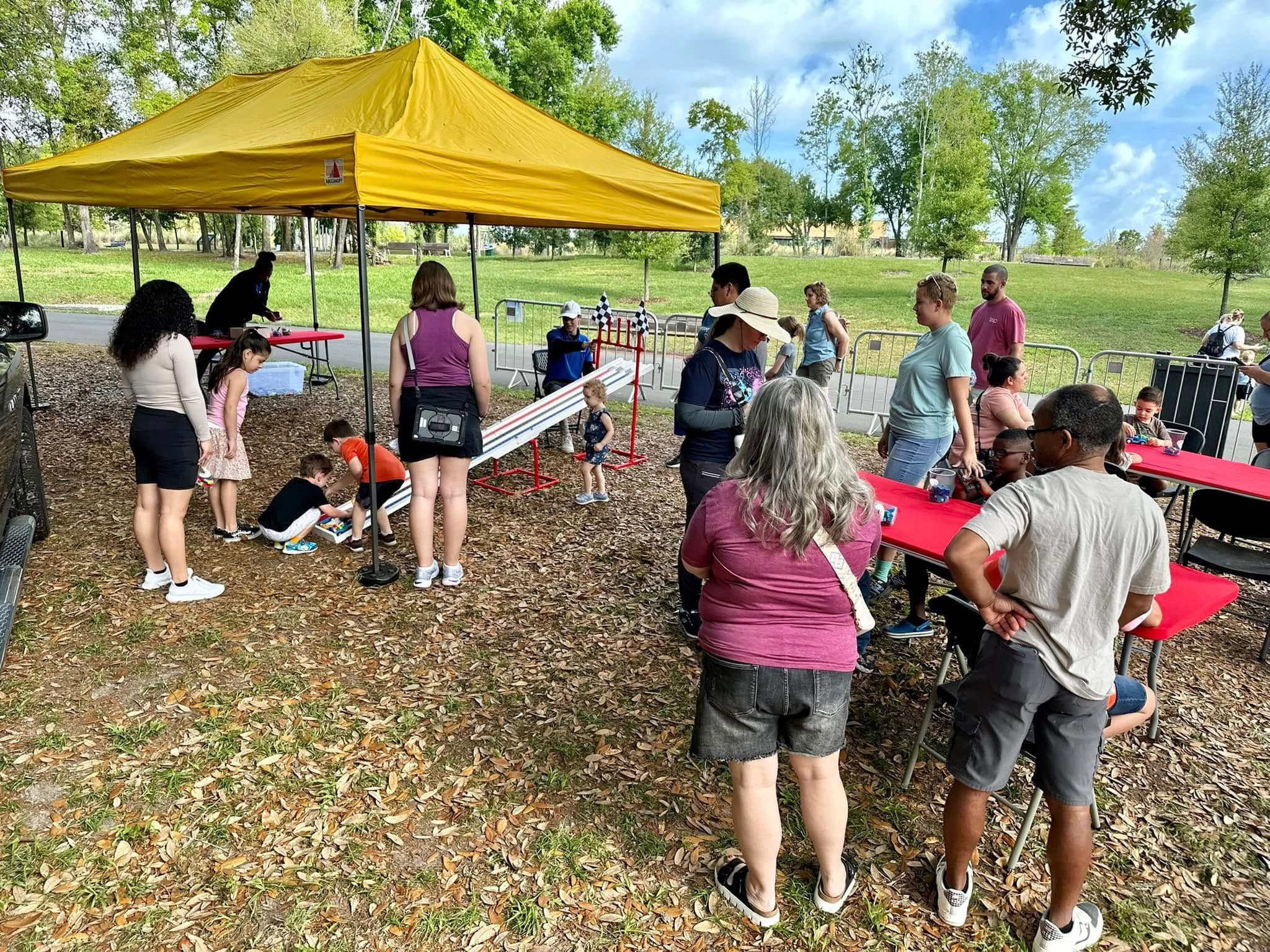 Families build at red tables and race cars on the Derby ramp under a yellow canopy in a park