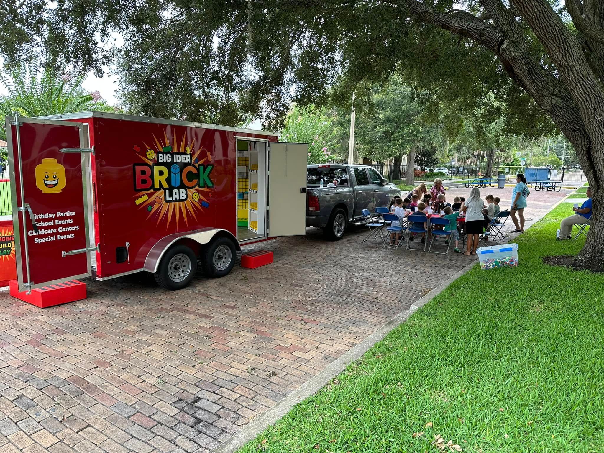 The Brick Lab trailer parked under oak trees while a group of kids builds at tables beside it