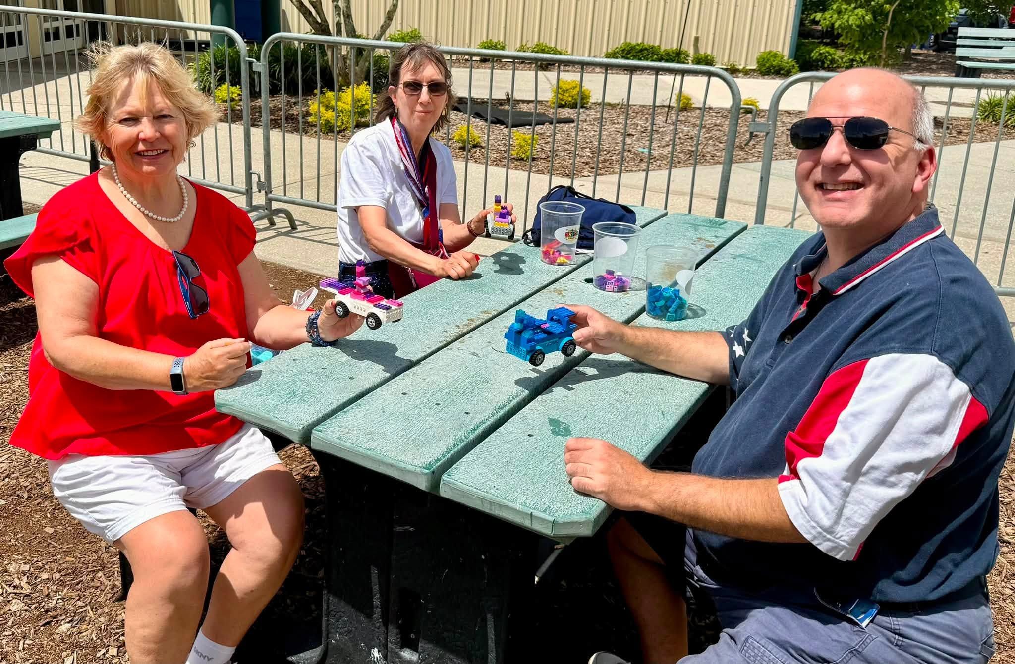 Three adults smile as they hold up the brick cars they built at a picnic table