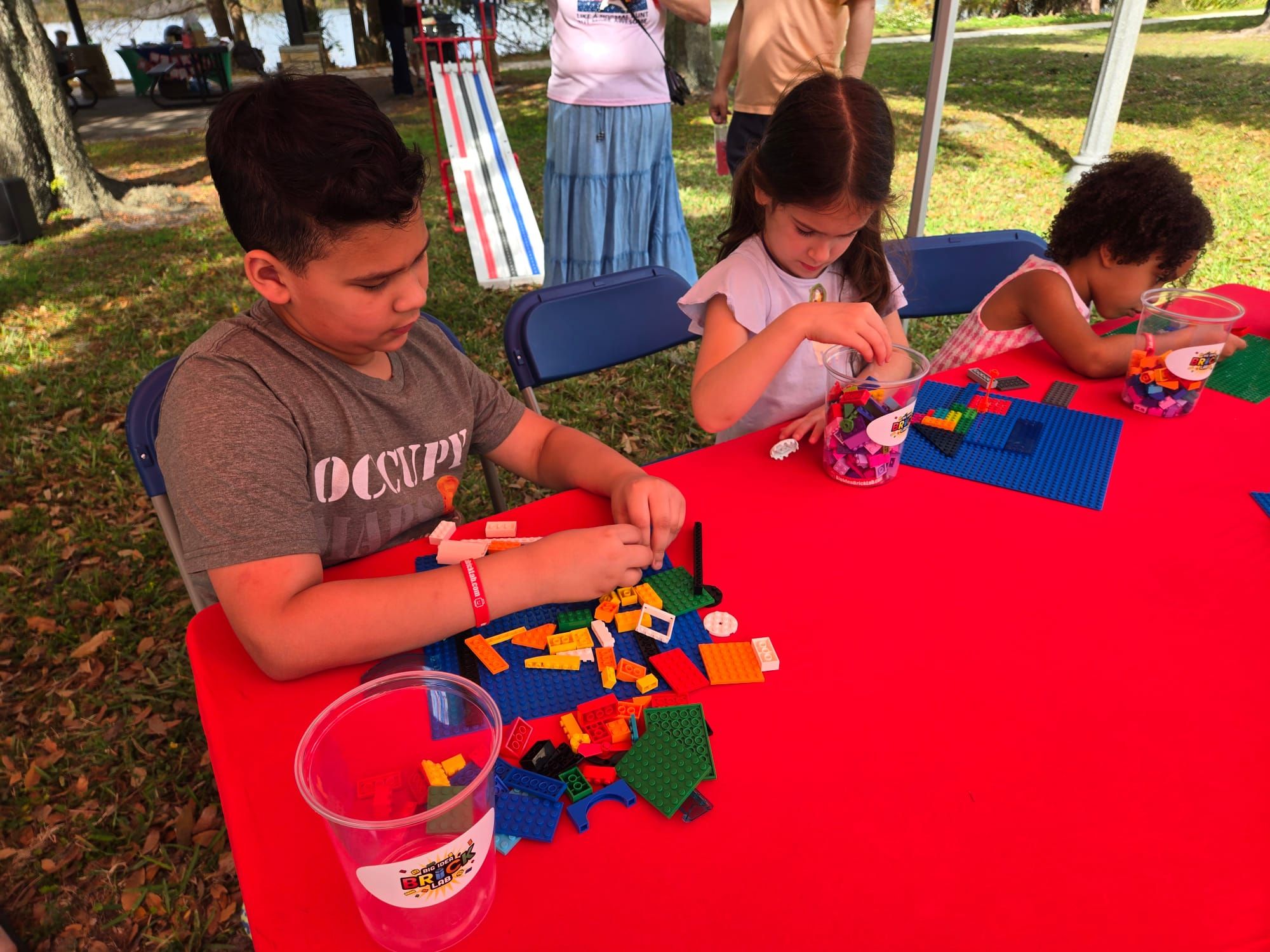 Three kids build on baseplates at a red table, with the Derby ramp set up on the grass behind
