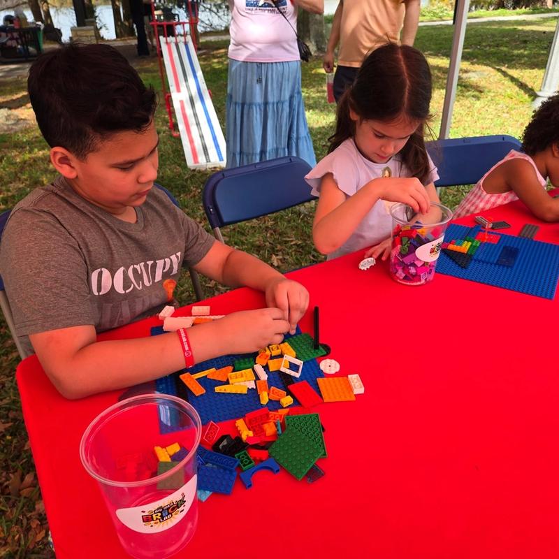 Three kids build on baseplates at a red table, with the Derby ramp set up on the grass behind