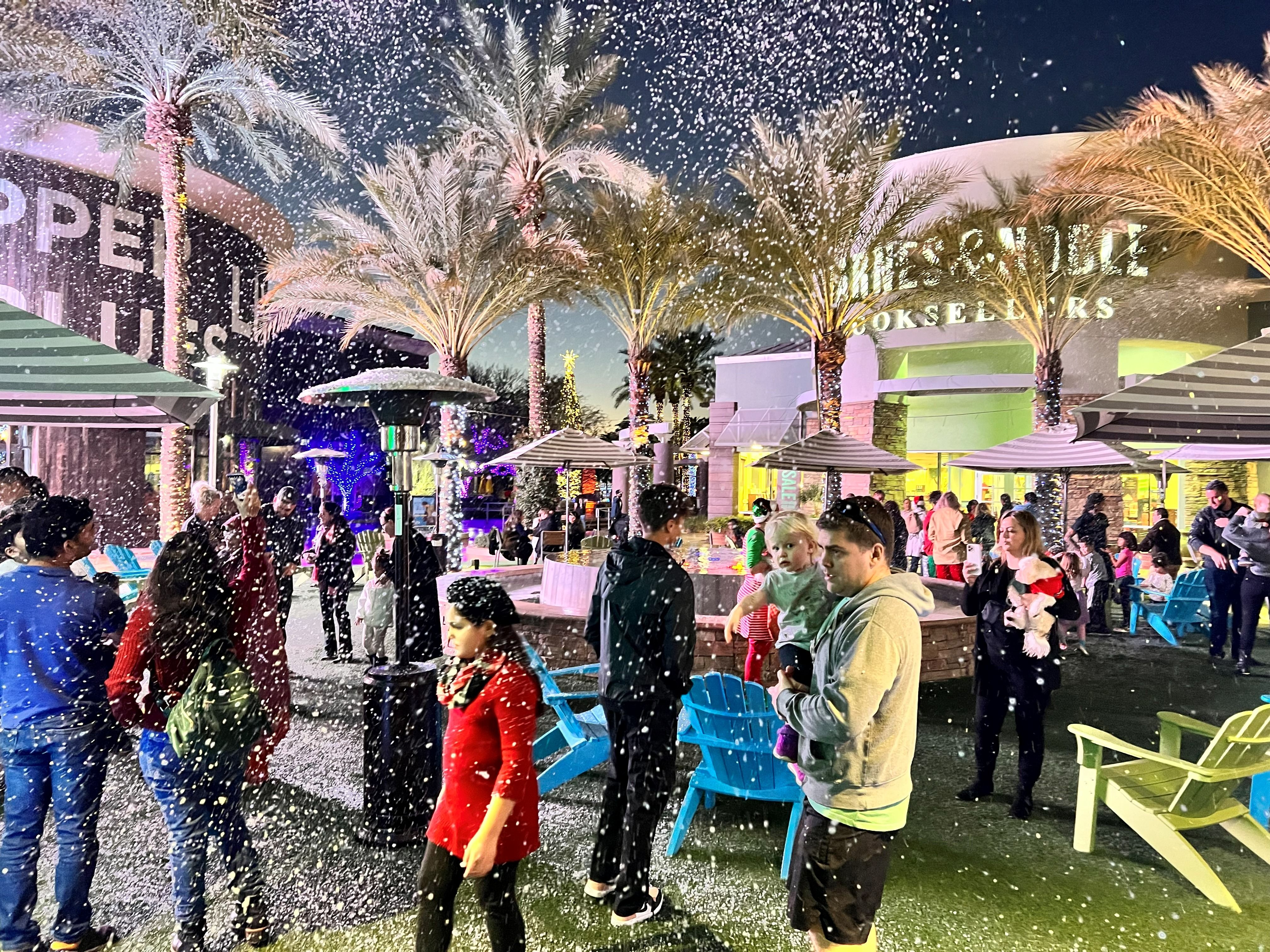 A parent holds a toddler as snow falls over a palm-lined plaza at a holiday night event