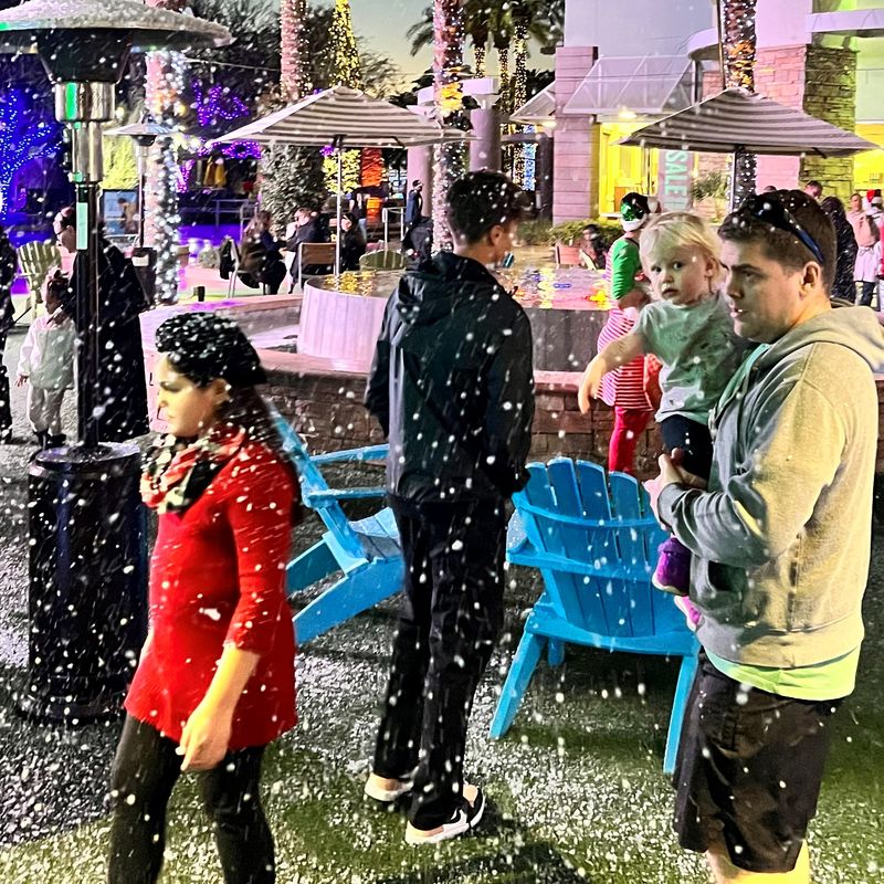 A parent holds a toddler as snow falls over a palm-lined plaza at a holiday night event