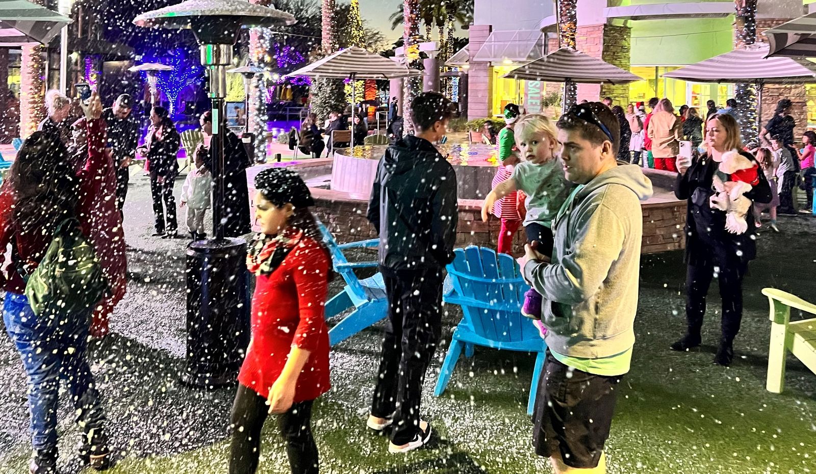 A parent holds a toddler as snow falls over a palm-lined plaza at a holiday night event