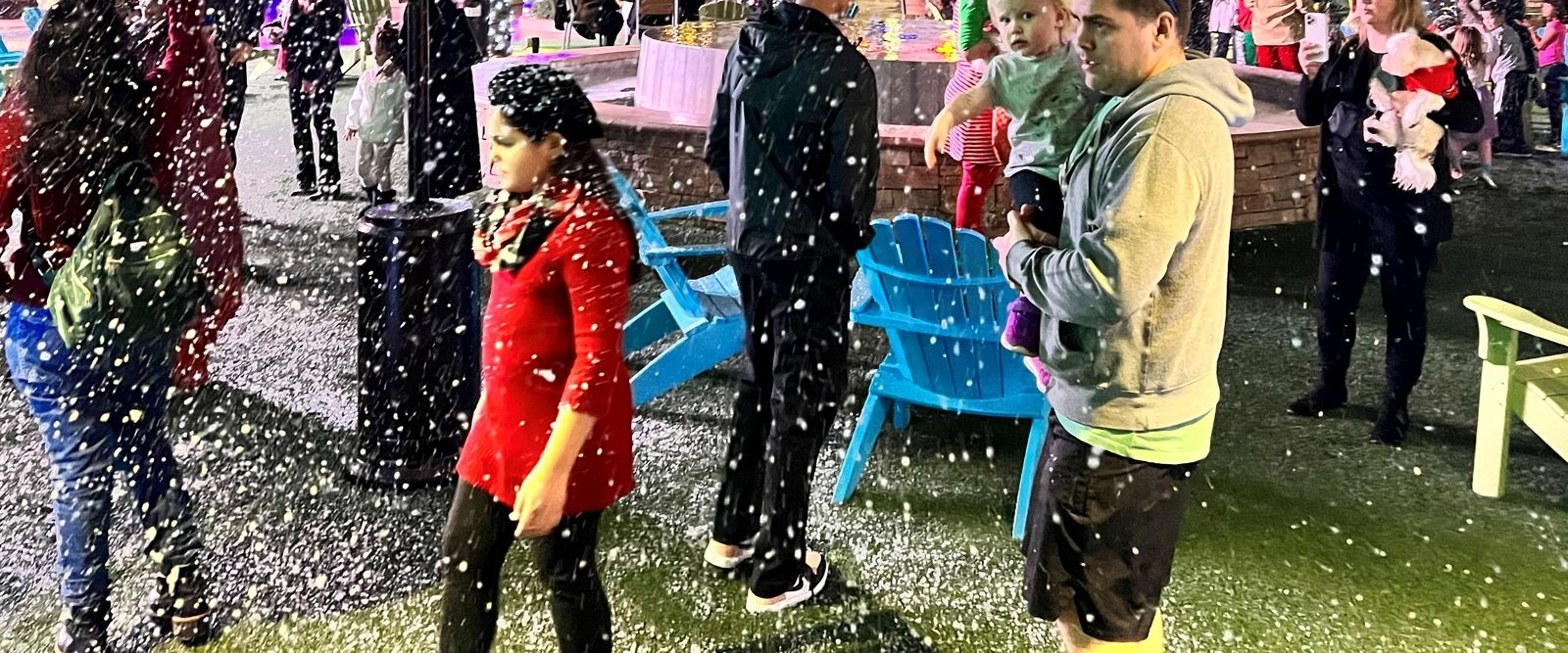 A parent holds a toddler as snow falls over a palm-lined plaza at a holiday night event