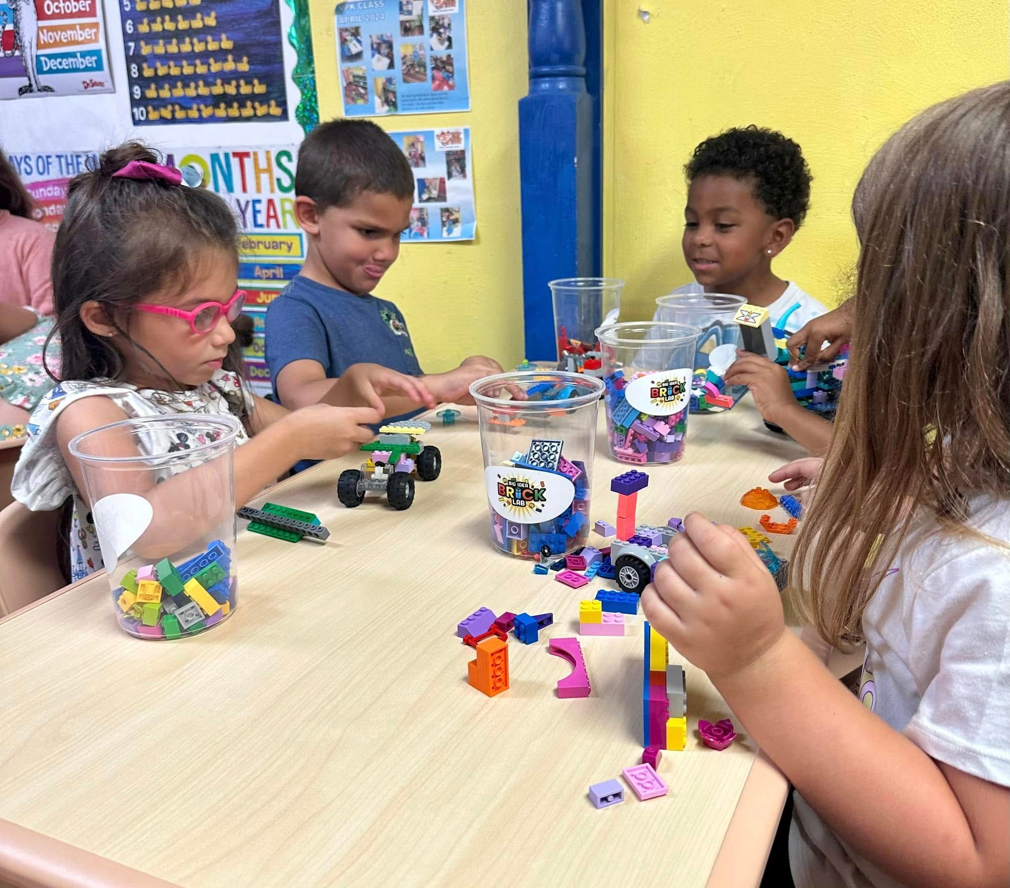 Preschoolers build brick cars at a classroom table from cups of building bricks