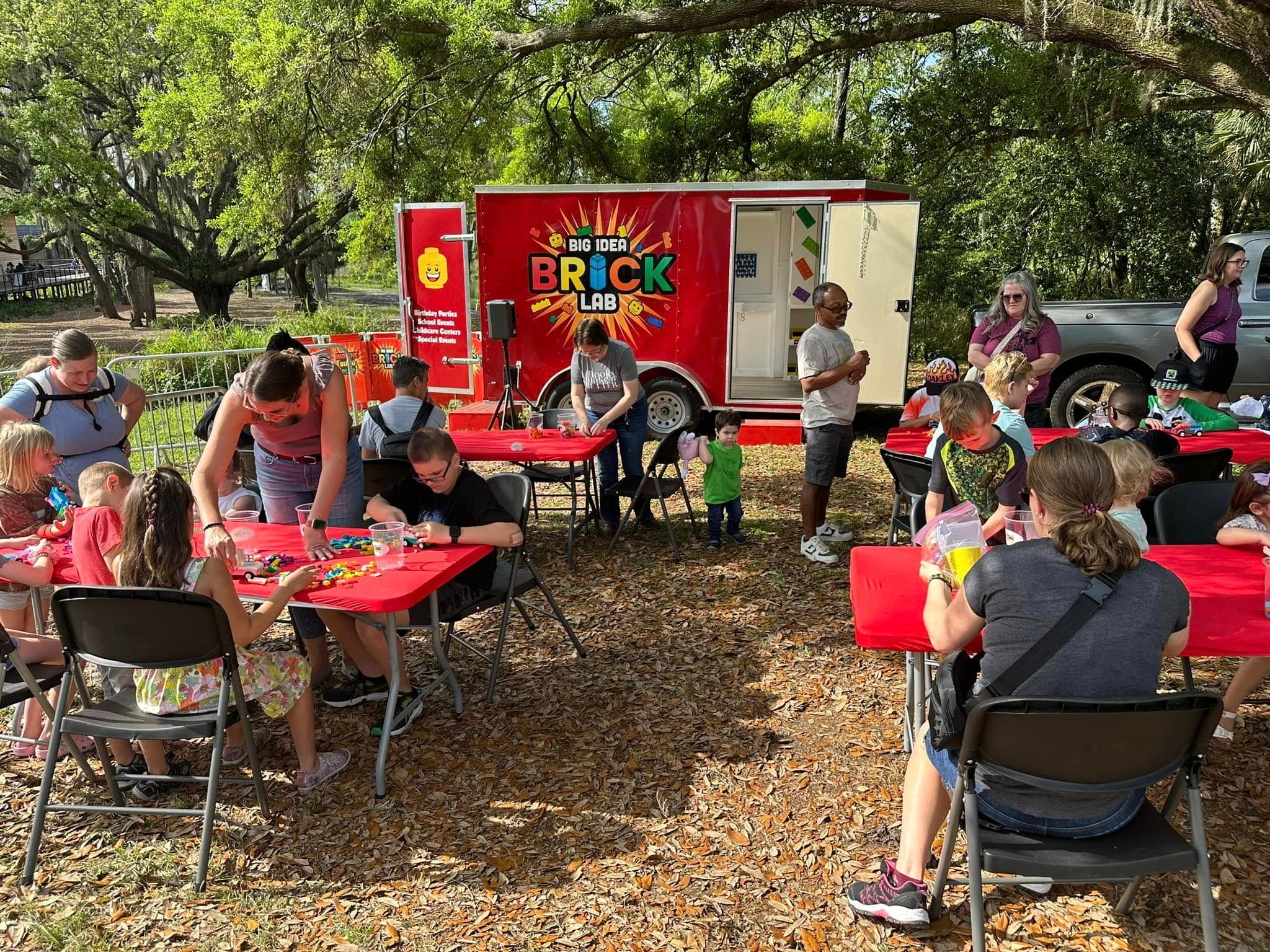 Families build at red tables in a shady park, with the Brick Lab trailer open behind them