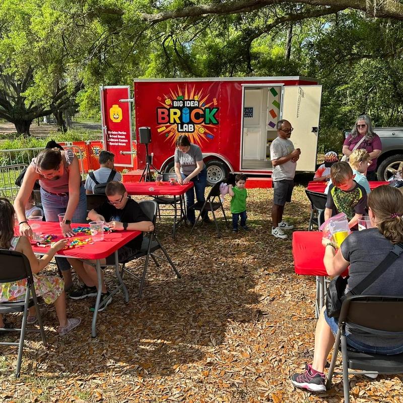 Families build at red tables in a shady park, with the Brick Lab trailer open behind them