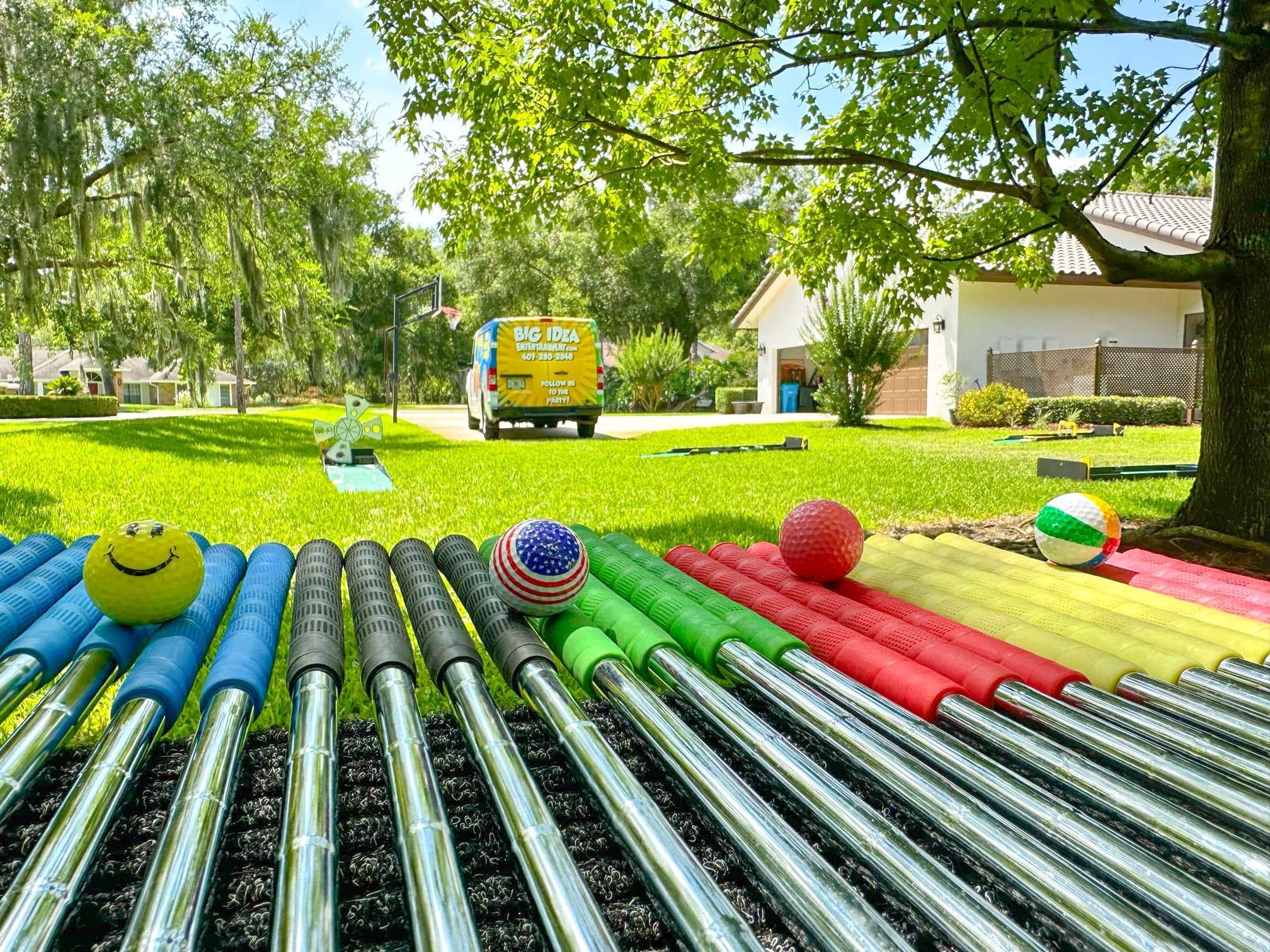 a rack of colored putters and balls in front of a mini golf hole on a shaded lawn