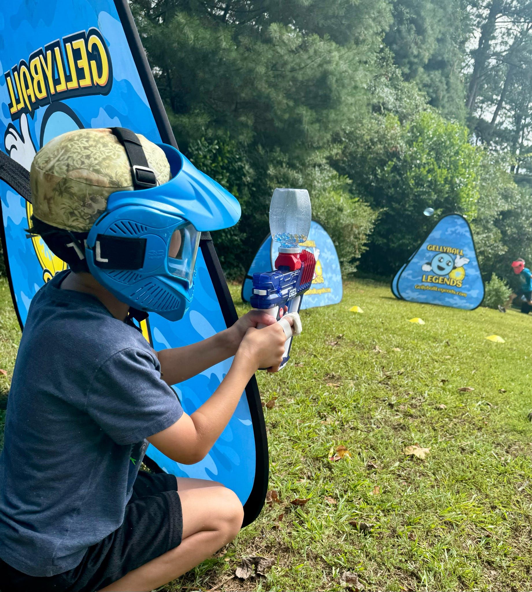 A boy in a camo cap and blue mask aims from behind a bunker toward the far side of the field