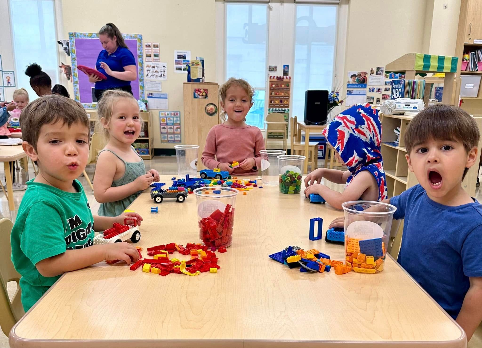 Young kids build brick cars at a classroom table with cups of red, green and orange bricks