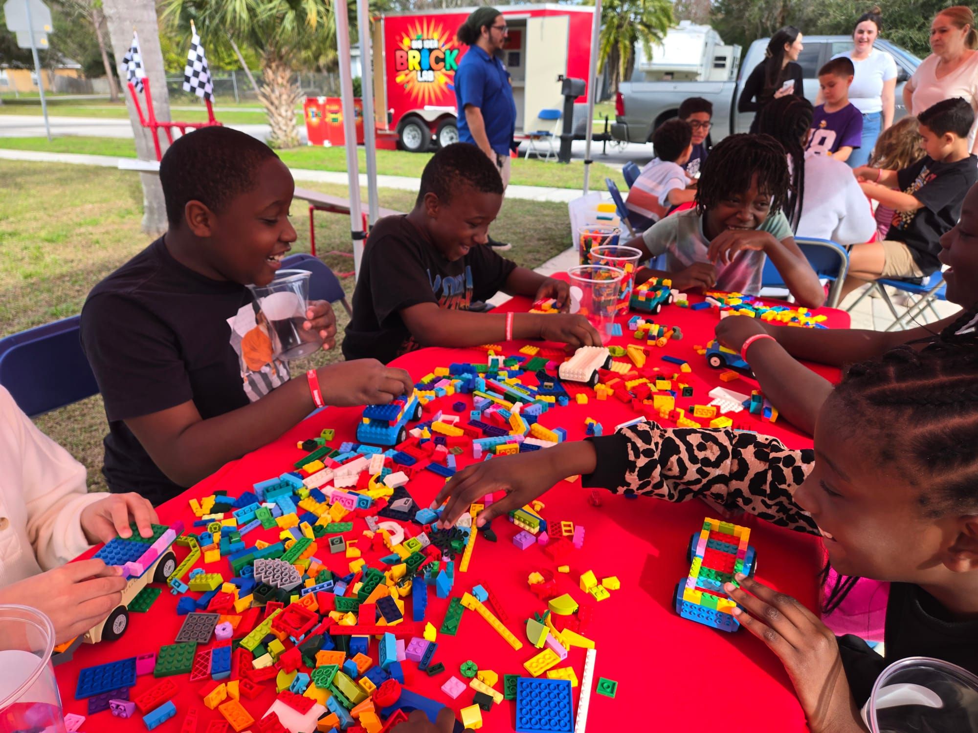 Kids laugh as they build brick cars at a red table, with the Brick Lab trailer parked behind
