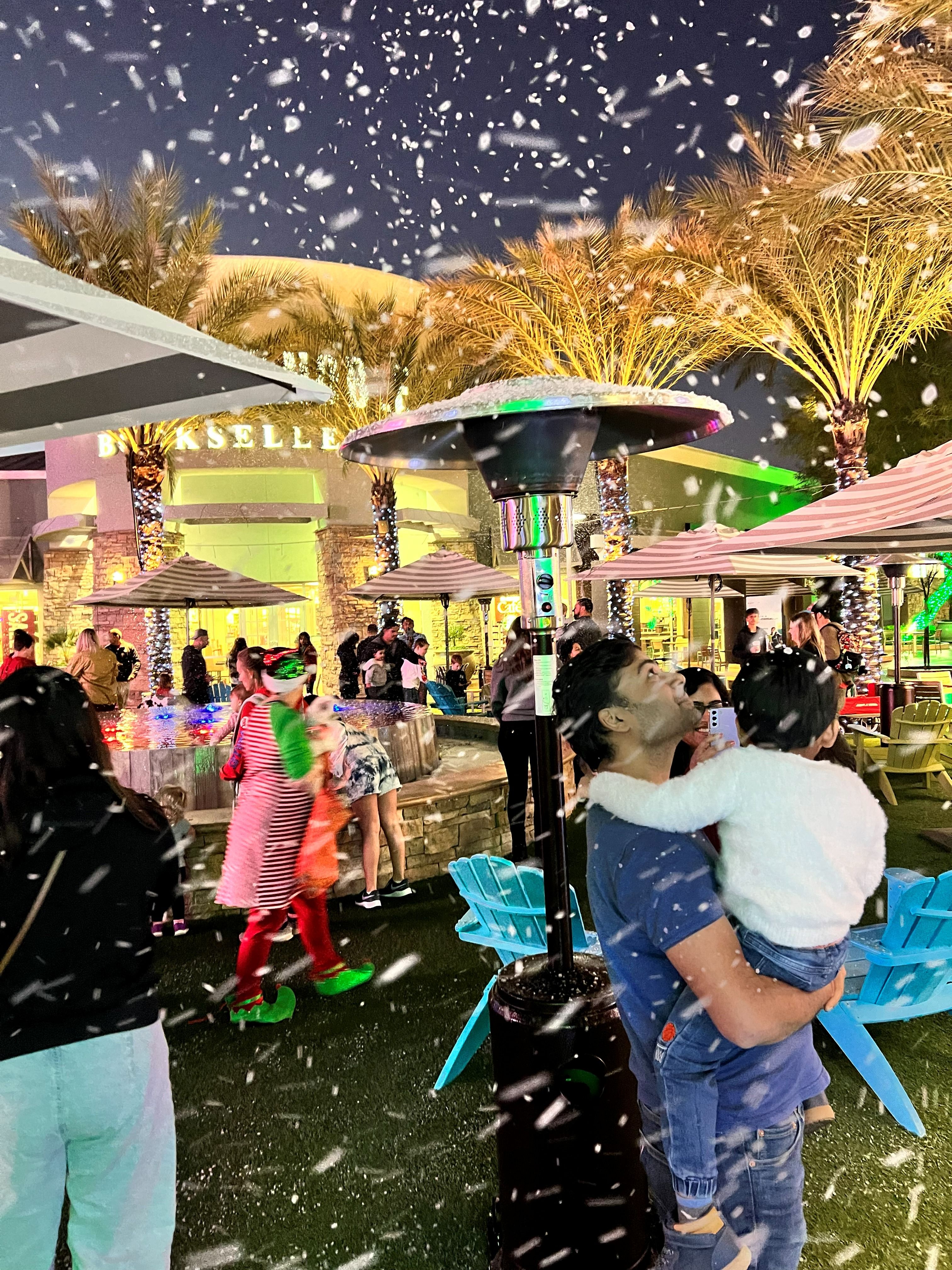 A child in a parent's arms looks up as snow falls at a holiday night event