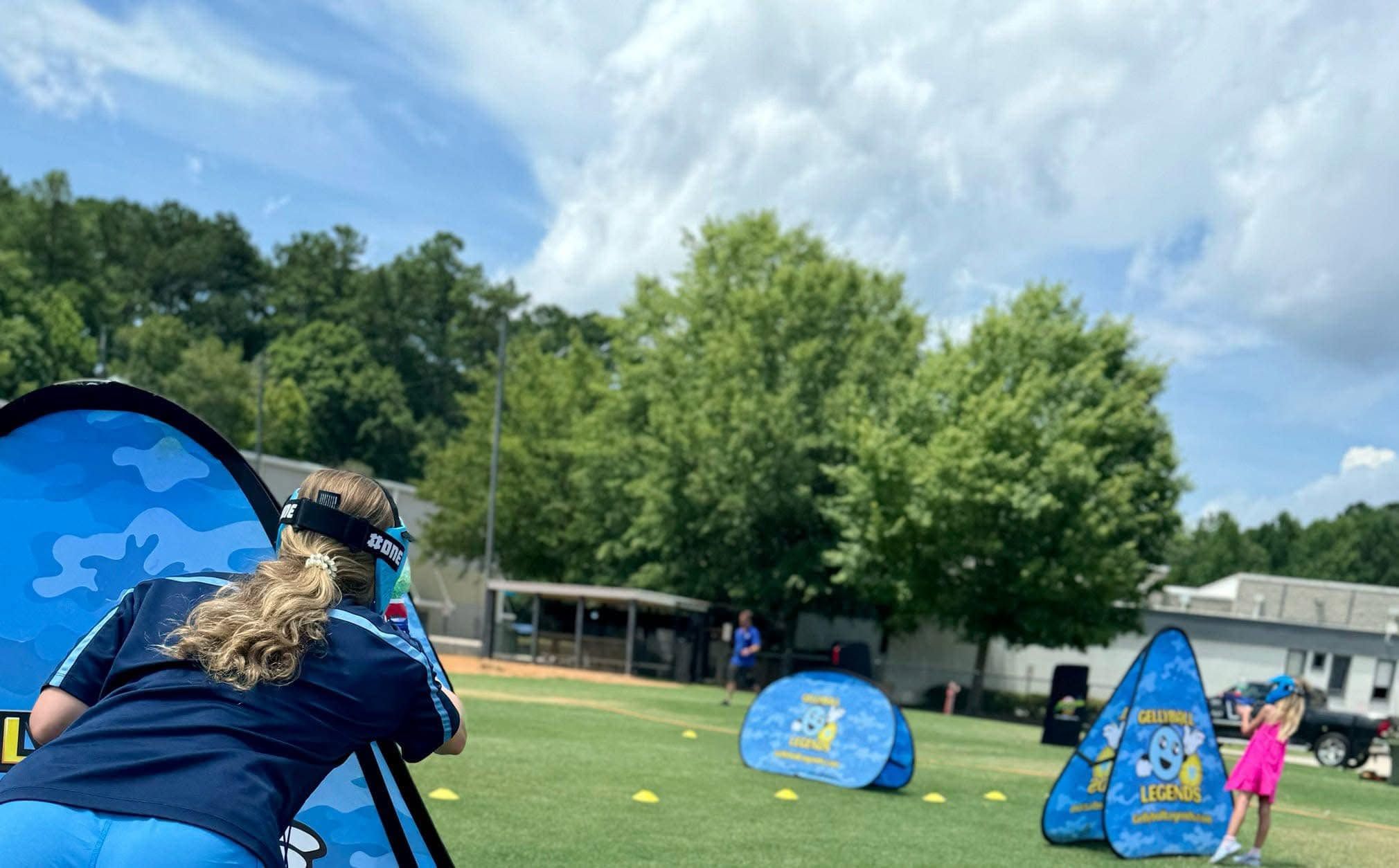 A player crouches behind a pop-up bunker while kids play gellyball across a sunny sports field