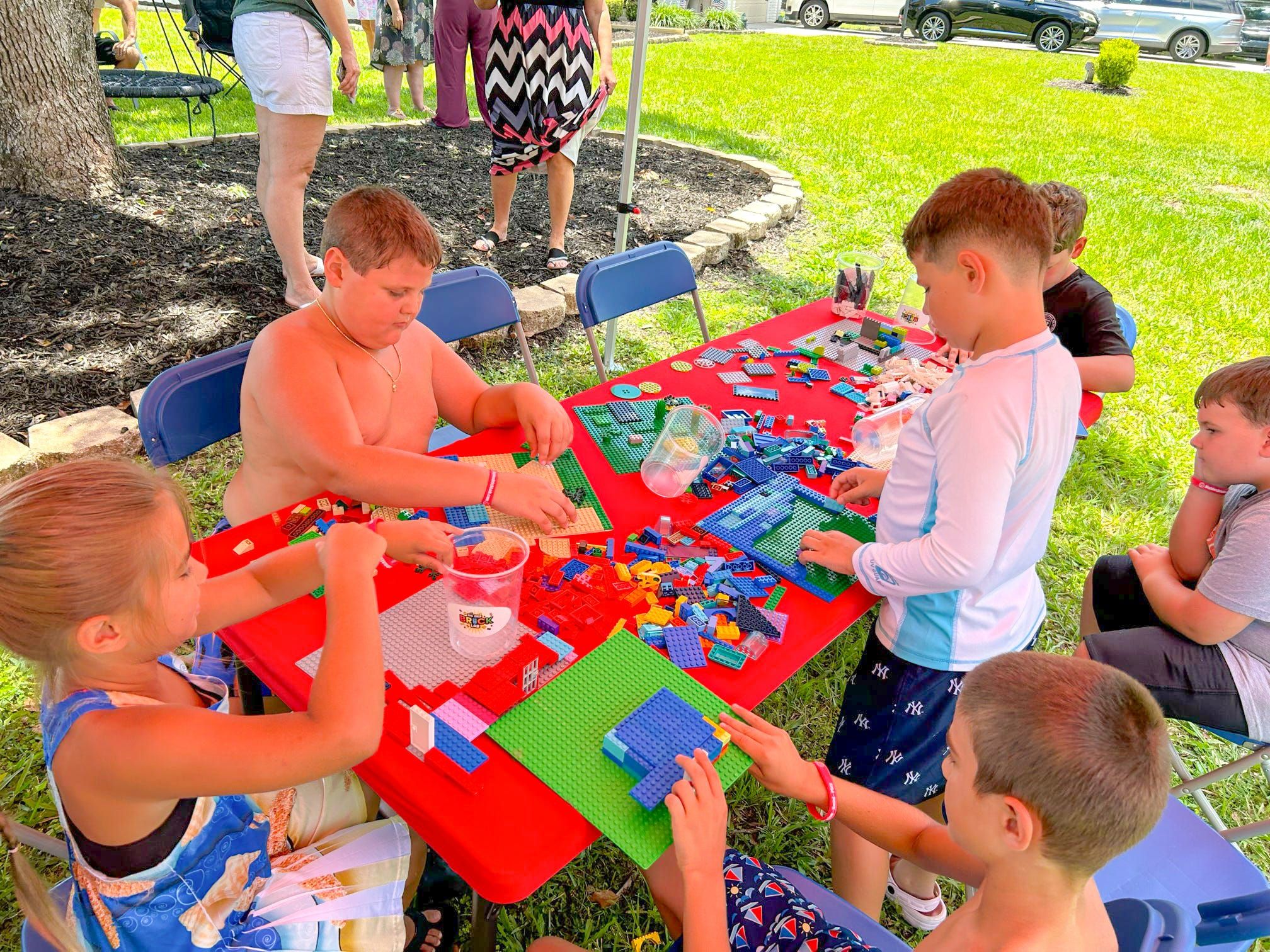 Kids build on baseplates around a red-covered table under a tent on a backyard lawn