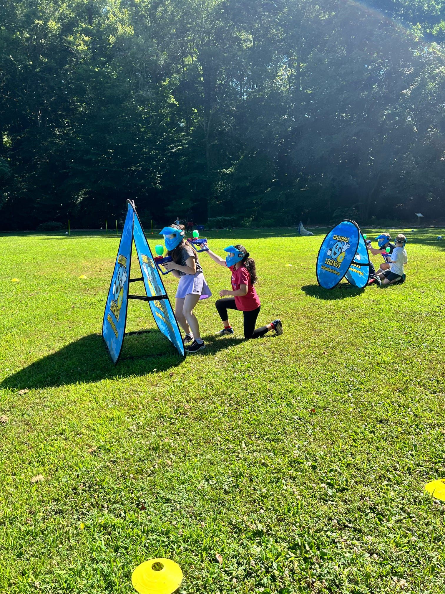 Two girls in blue masks take aim from an A-frame bunker on a sunlit field