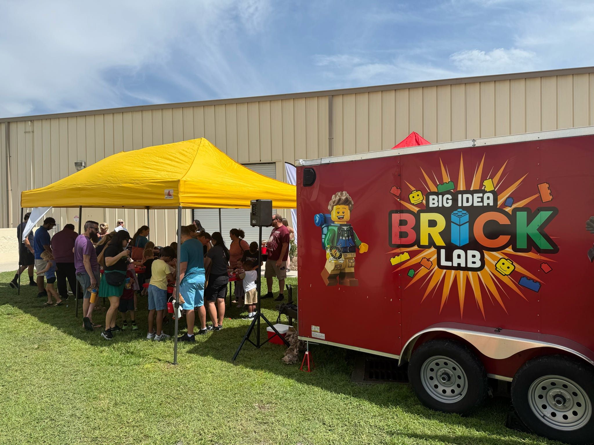 Families crowd a build table under a yellow canopy beside the Big Idea Brick Lab trailer at an outdoor event