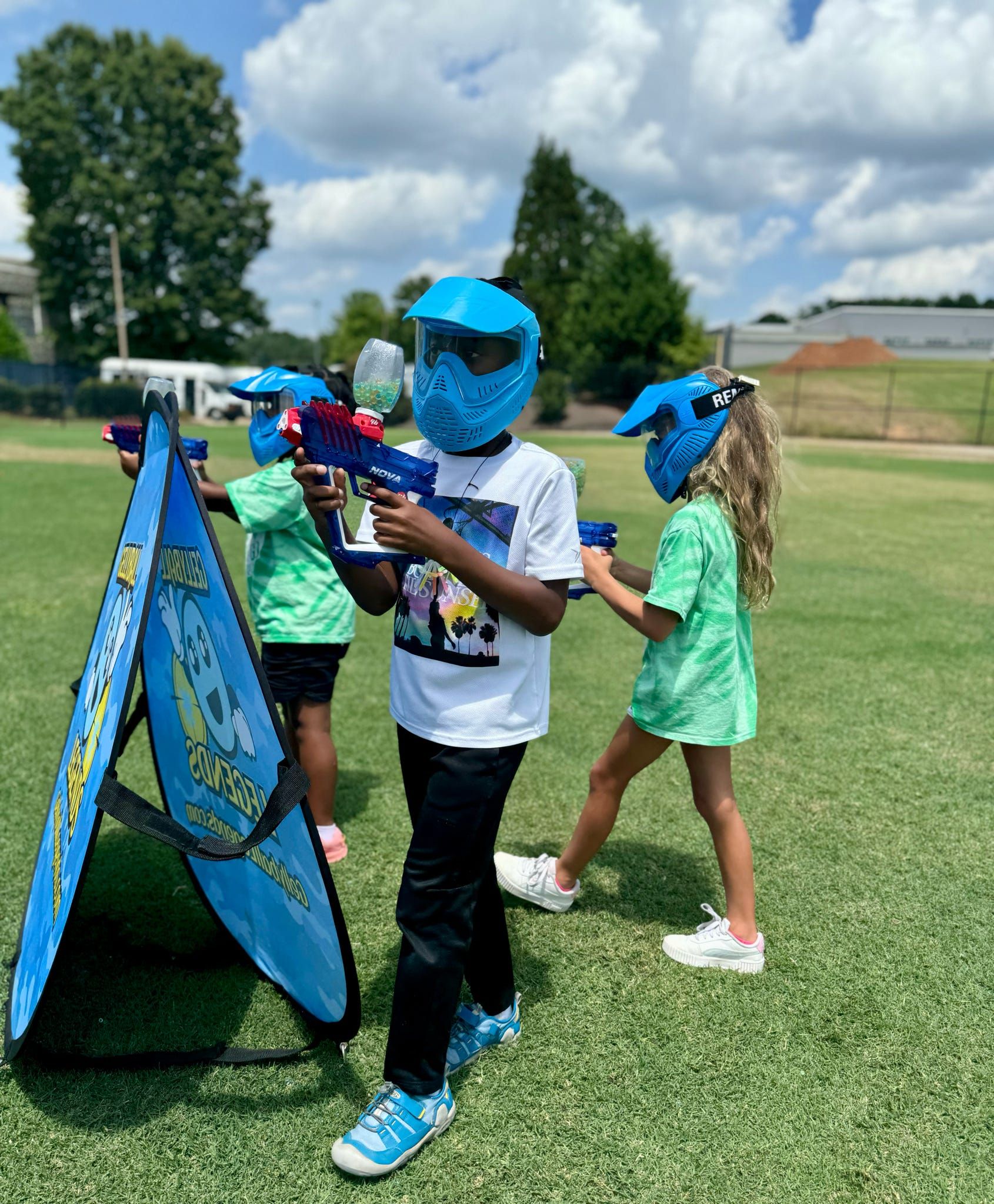 Three kids in blue masks play gellyball beside a pop-up bunker on a sports field