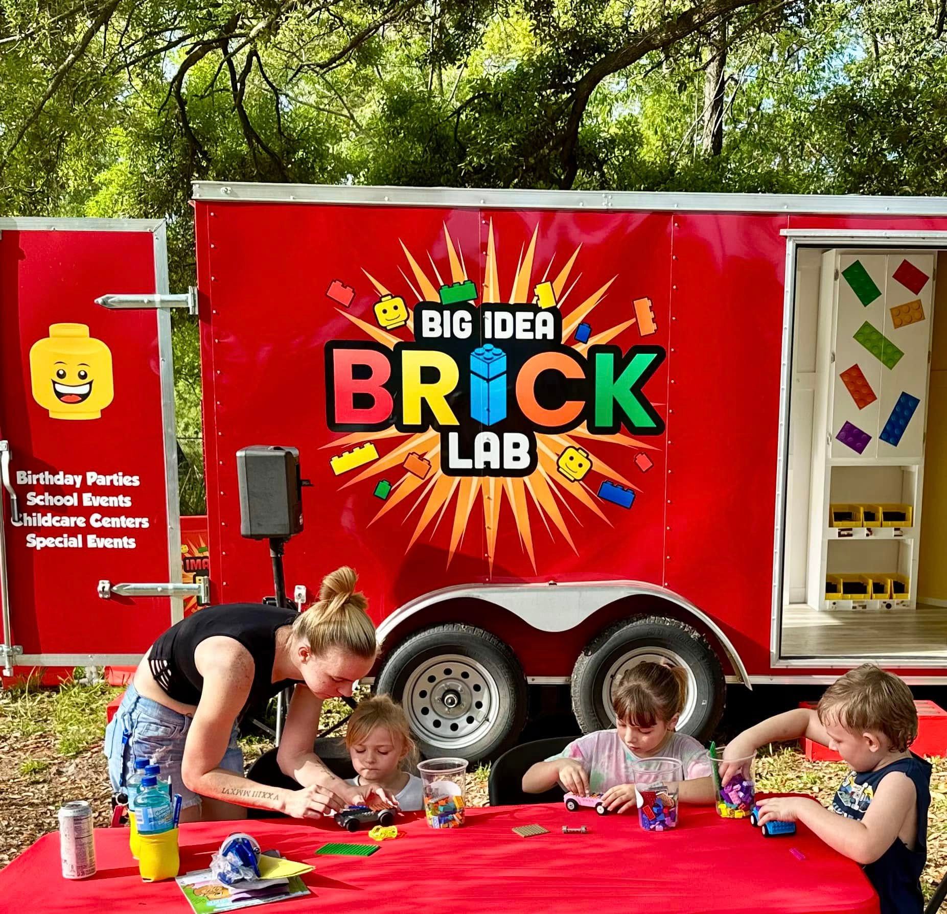 An adult helps three kids build brick cars at a red table in front of the Brick Lab trailer