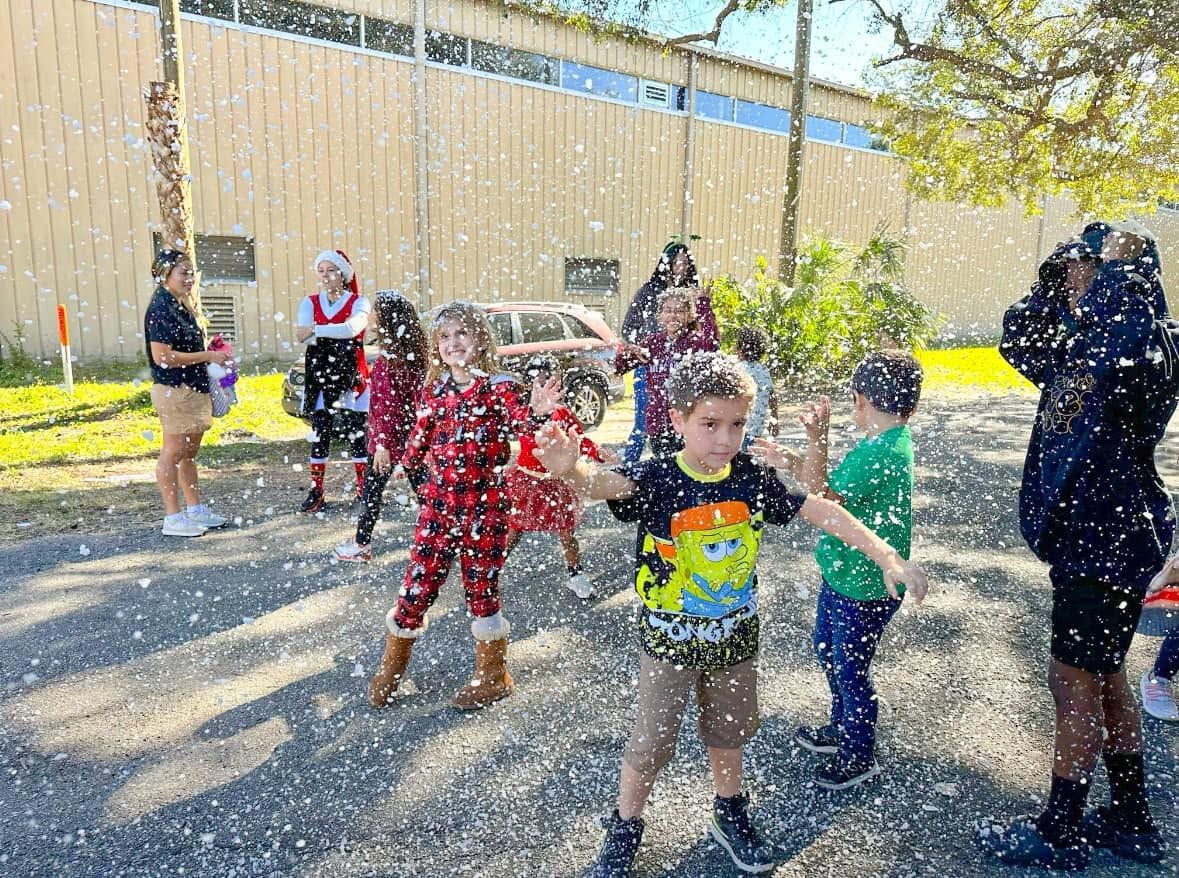 Kids in holiday pajamas play in falling snow beside a costumed holiday helper