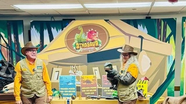 Two Dinosaur Adventures presenters at the program set with geologic period signage and a dinosaur puppet