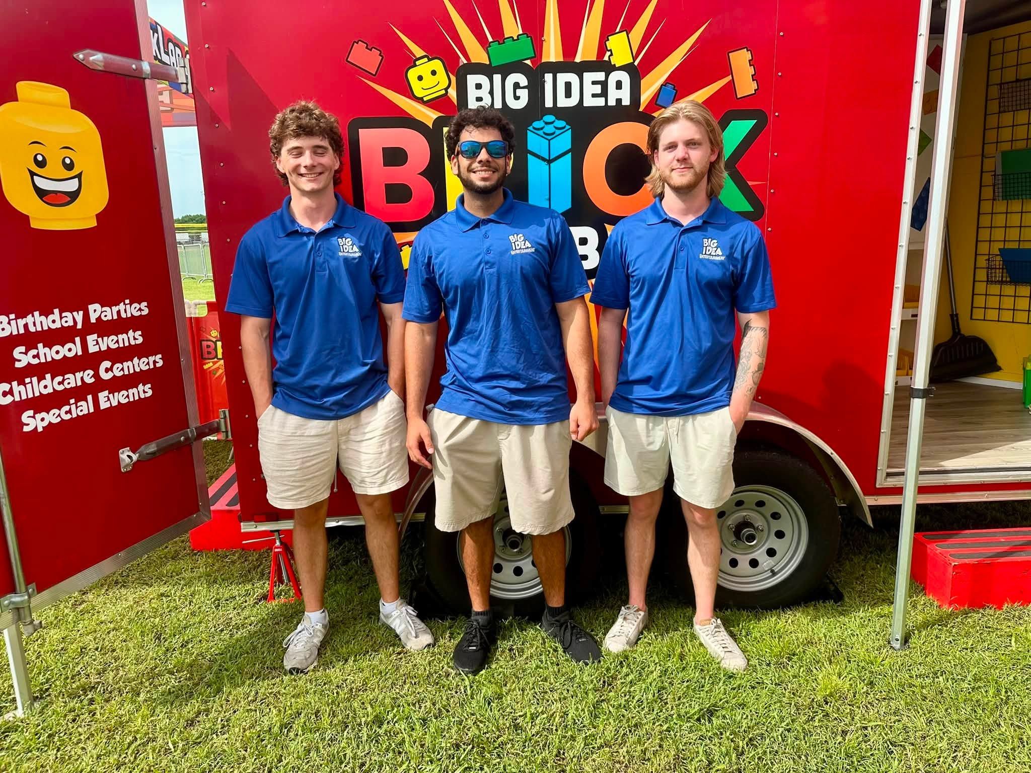Three crew members in matching blue polos stand in front of the Big Idea Brick Lab trailer