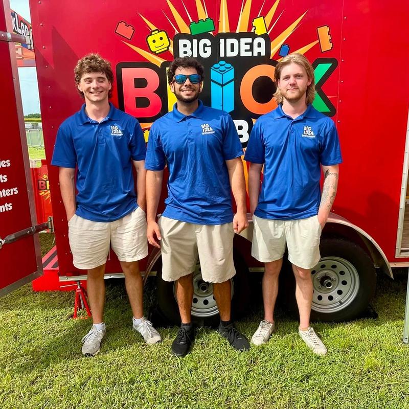 Three crew members in matching blue polos stand in front of the Big Idea Brick Lab trailer