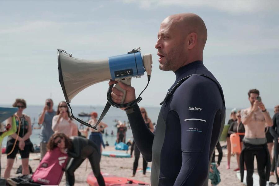 Hugo Tahholm, CEO of SAS, is pictured from the waist up in a wetsuit. He holds a megaphone and speaks through it to assembled protestors on the beach, who can be seen in the background.
