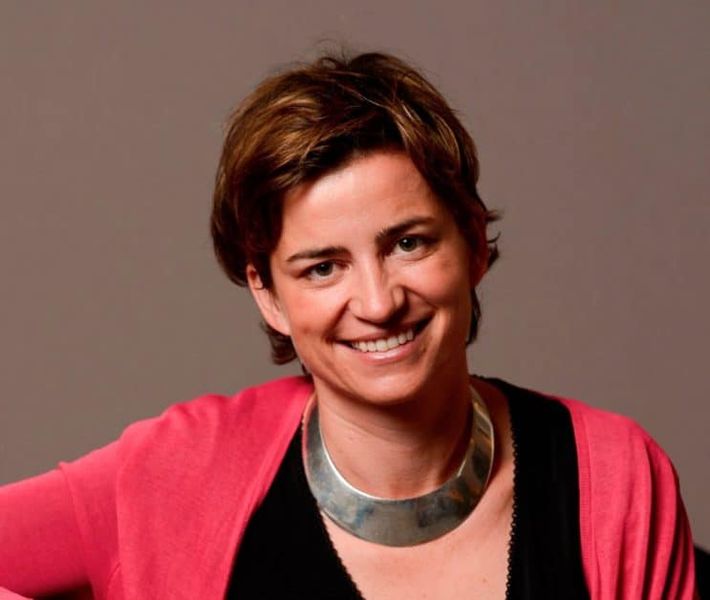 Headshot of Sophie Lambin smiling, looking directly into the camera against a neutral background. She wears a coral cardigan, black top and chunky silver necklace. Her hair is short and brown, with gold highlights,